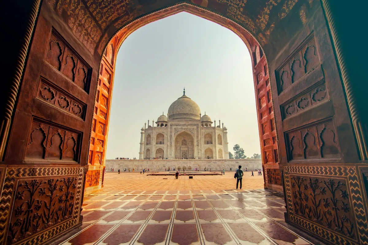 The Taj Mahal seen through a carved red sandstone archway, with visitors on the courtyard