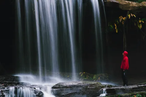 A hiker in a red hoodie looks up at a forest waterfall near Aizawl, Mizoram