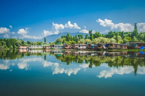 Houseboats along the shore of a still lake in Srinagar, mirrored in the water under a blue sky